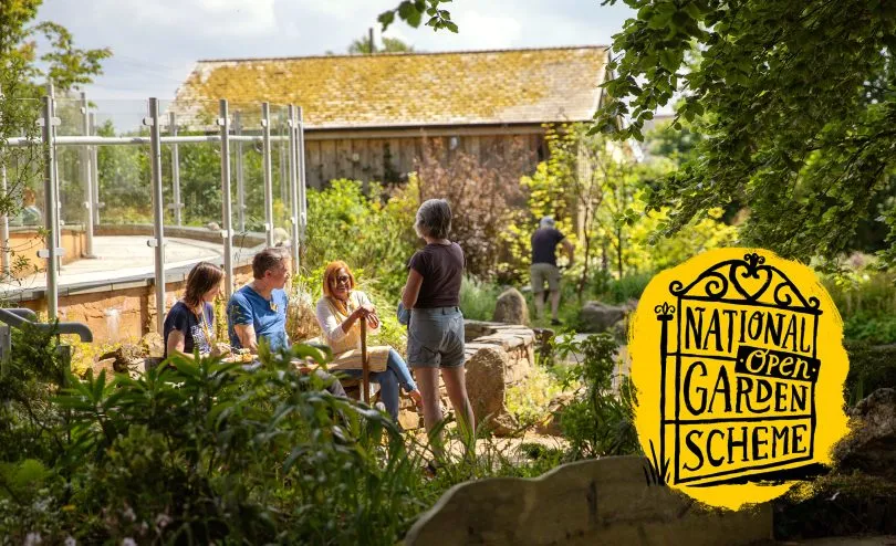 Four people talking in a sunlit garden, three seated on stone among dense green planting, with a slate-roofed barn behind.