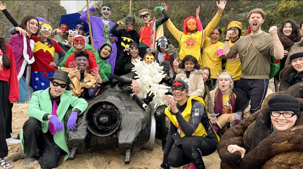 A crowd in superhero fancy dress packed around a home-made black car sculpture on the sand, arms raised.