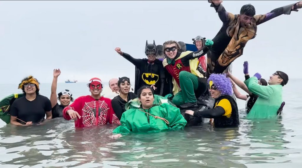 Swimmers in superhero costumes standing waist-deep in flat grey sea, one leaping in at the right.