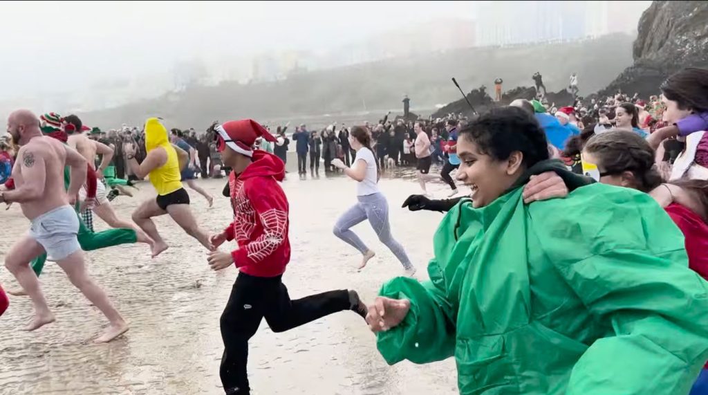Swimmers in Christmas hats and fancy dress running across wet sand into the sea, spectators lining the shore.