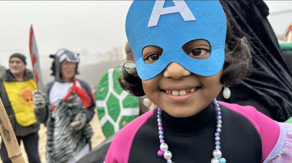 A smiling girl in a blue Captain America mask and pink wetsuit, with costumed swimmers blurred behind her.